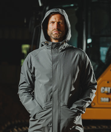 Man wearing a grey Caterpillar jacket, with the hood up, hand in pockets and machinery behind him in a dark building