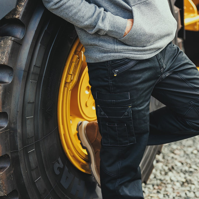 Close-up image of a man leading against a large plant machine tyre, wearing a pair of black Caterpillar Operator FX Trousers